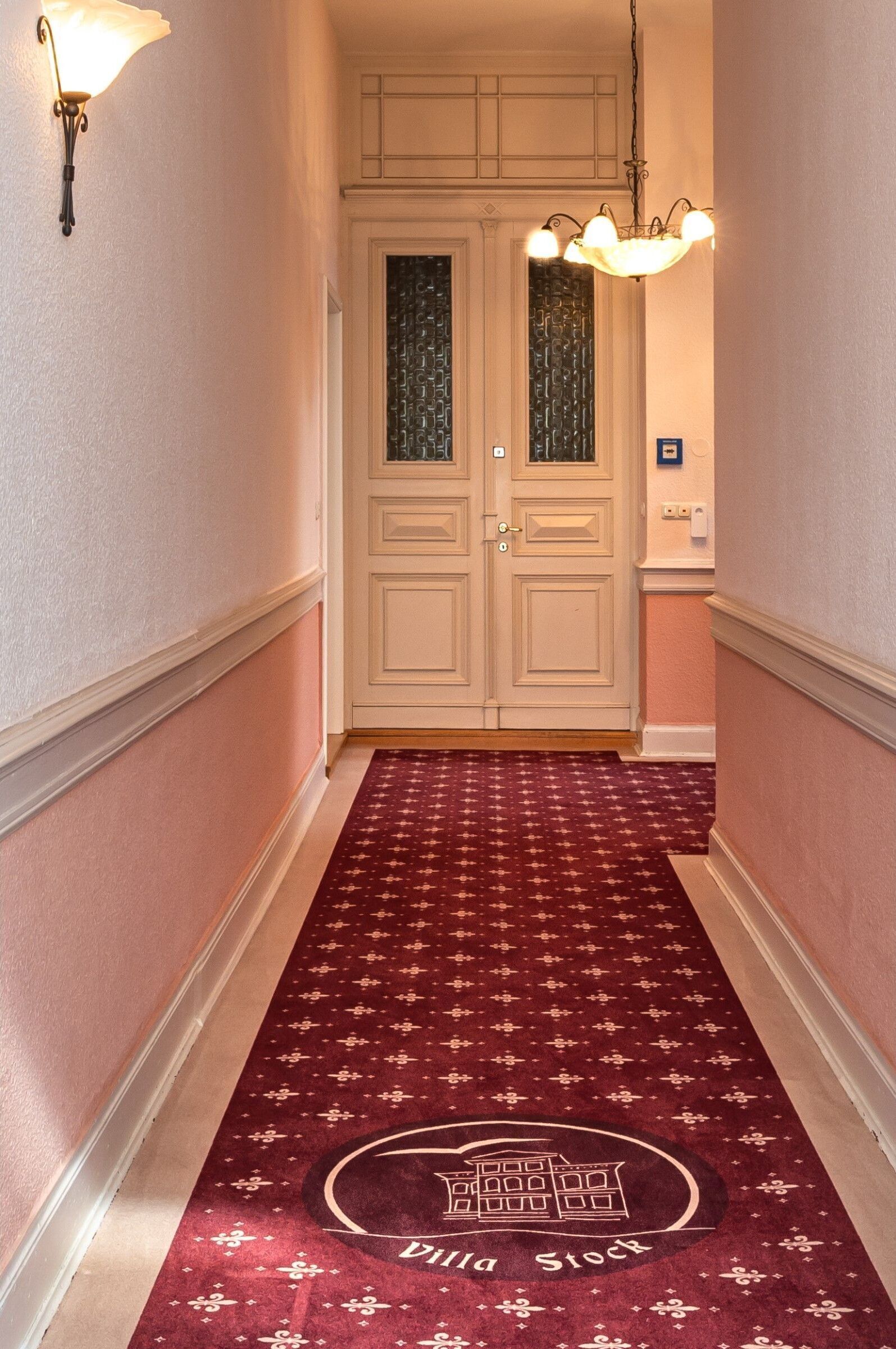 Un pasillo con alfombra roja que presenta el logotipo de un edificio, puertas dobles blancas y luces fijadas a la pared.