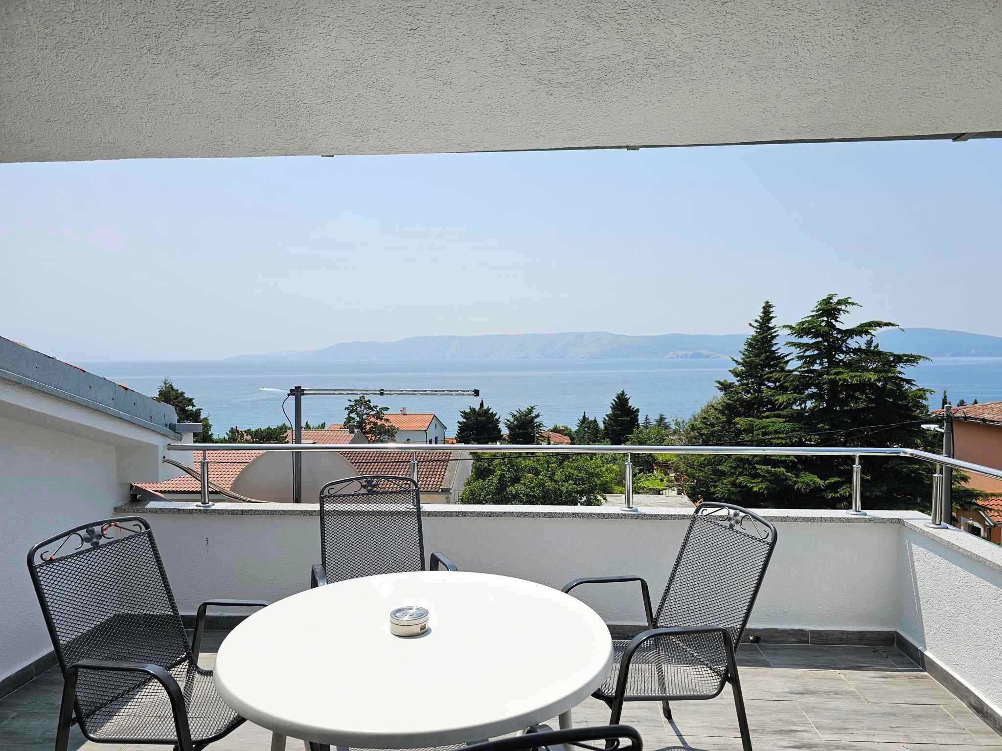 En balkon med et rundt bord og fire stole, med udsigt over et hav og fjerne bjerge.