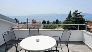 Un balcon avec une table ronde et quatre chaises, donnant sur une mer où l’on aperçoit des montagnes au loin.