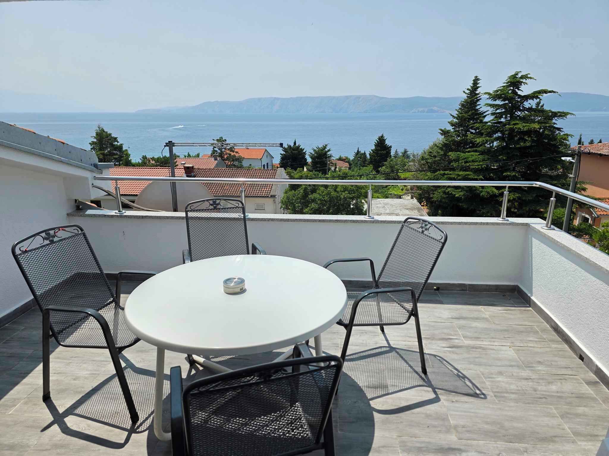 Une table ronde entourée de quatre chaises, sur un balcon donnant sur une étendue d’eau et des montagnes lointaines.