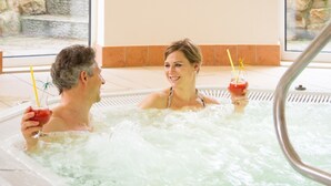 A couple in a hot tub with drinks, surrounded by stone walls and a tiled floor. - Deluxe Double Room - Hotel Teutoburg Forest GmbH (Tecklenburg)