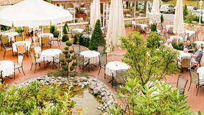 An outdoor patio area with round tables and chairs, surrounded by greenery and a small pond. - Deluxe Double Room - Hotel Teutoburg Forest GmbH (Tecklenburg)