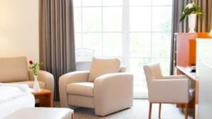 A hotel room with a bed, two armchairs, a desk, and a large window with curtains. - Deluxe Double Room - Hotel Teutoburg Forest GmbH (Tecklenburg)