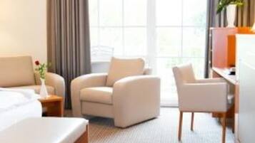Quarto de hotel com uma cama, duas poltronas, uma escrivaninha e uma janela grande com cortinas.