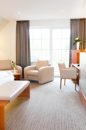 A hotel room with a bed, two armchairs, a desk, and a large window with curtains.