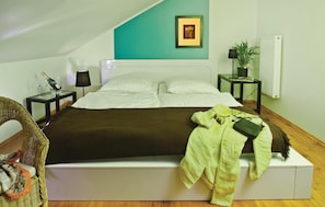 A neatly made bed with a white headboard, a green blanket, and a green jacket on the footboard. There is a wicker chair with a colorful scarf, a small table with a lamp, and a framed picture on the wall. - 2 bedroom amazing home in Pinnow (Pinnow)