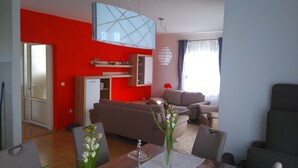 A modern living room with a red accent wall, a sofa, a dining table with a vase of flowers, and a kitchen area with a fridge and cabinets.