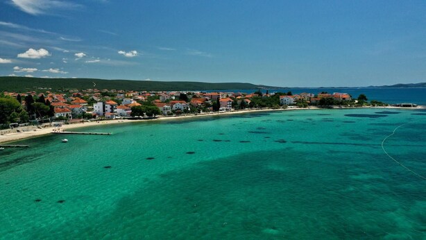 A coastal town with clear turquoise waters, a sandy beach, and a row of buildings with red roofs.
