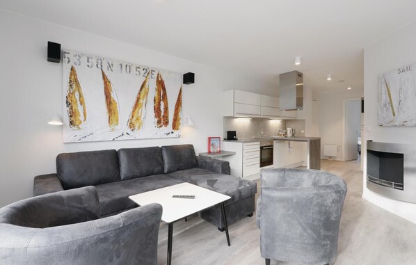 A modern living room with a grey sofa, a white coffee table, and a grey armchair. There is a kitchen area with white cabinets and a built-in oven. A large painting with abstract shapes is on the wall, and a fireplace is visible in the background.