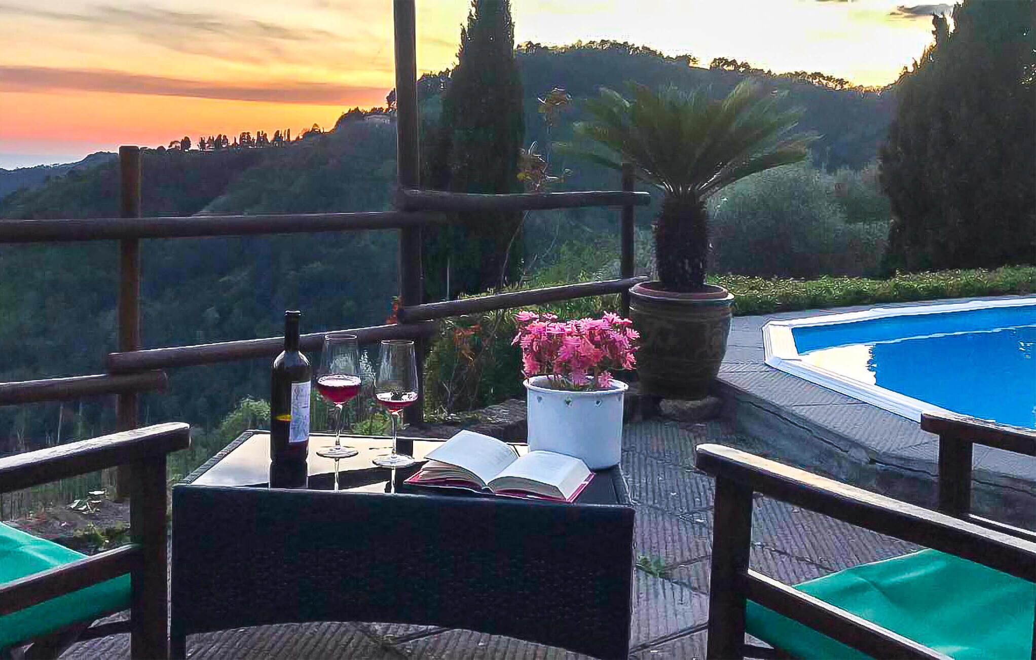 A terrace with a view of a mountainous landscape, a swimming pool, and a table with a book, wine glasses, and a bottle.