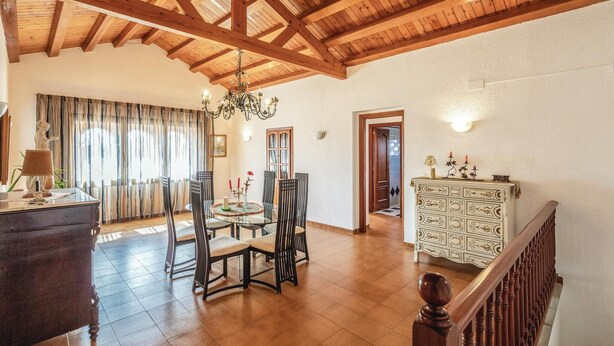 Een ruime eetruimte met een houten plafond en een kroonluchter, een eettafel met stoelen en een wit kastje met decoratieve objecten.