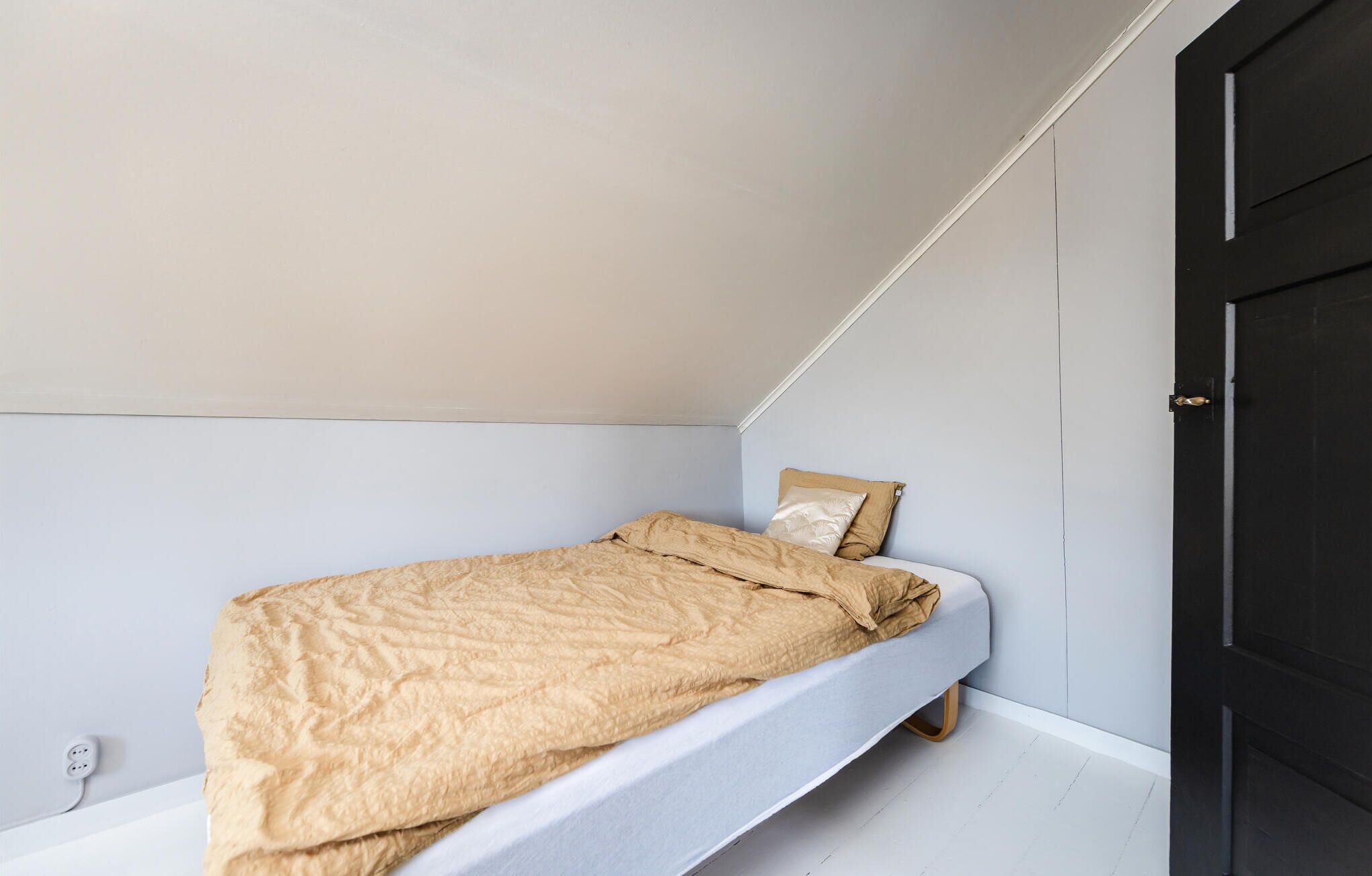 Una habitación pequeña con cama individual, cabecera blanca, manta color beige y almohada.