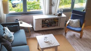 Ett mysigt vardagsrum med en blå soffa, ett litet vitt bord och en blå stol. Ett fönster med gardiner syns, och en växt står på en hylla.
