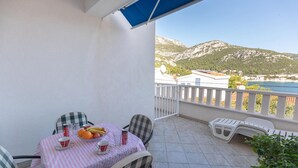 En balkon med et bord dækket til to, med skakternede stole og en pink-stribet dug. Der er en skål med frugt og røde kopper på bordet. Balkonen har udsigt over et bjergrigt landskab med en klar himmel.
