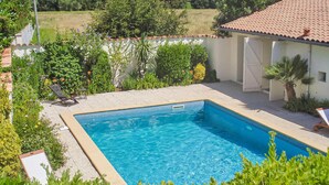 A rectangular swimming pool with a tiled deck surrounding it, set in a landscaped garden with various plants and a white building with a red-tiled roof in the background. - Stunning home in Cézac (Cézac)