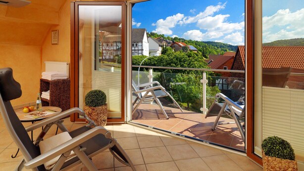 A balcony with a view of houses and trees, a chair, a small table with a bottle and fruit, and a potted plant. - Room type A - Double room - Flair Hotel Werbetal (Waldeck)