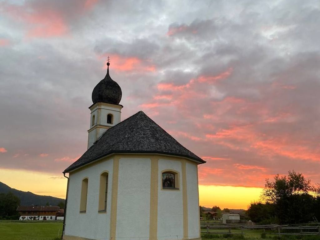 En lille kirke med et klokketårn og en kuppel, omgivet af en græsplæne og træer, under en solnedgangshimmel.