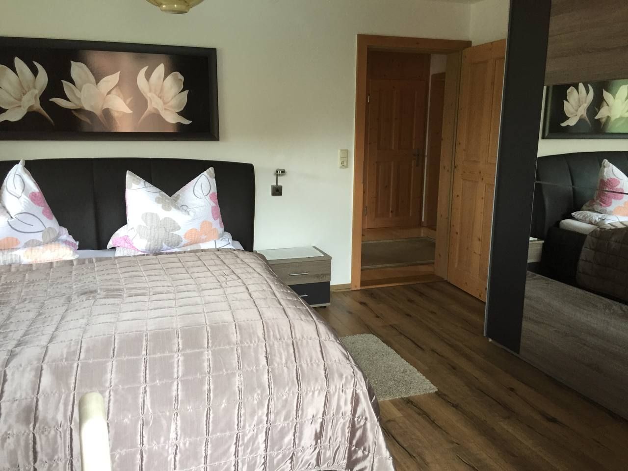 Ein Schlafzimmer mit einem großen Bett, Holzboden, einem Nachttisch und einer Tür zu einem weiteren Raum.