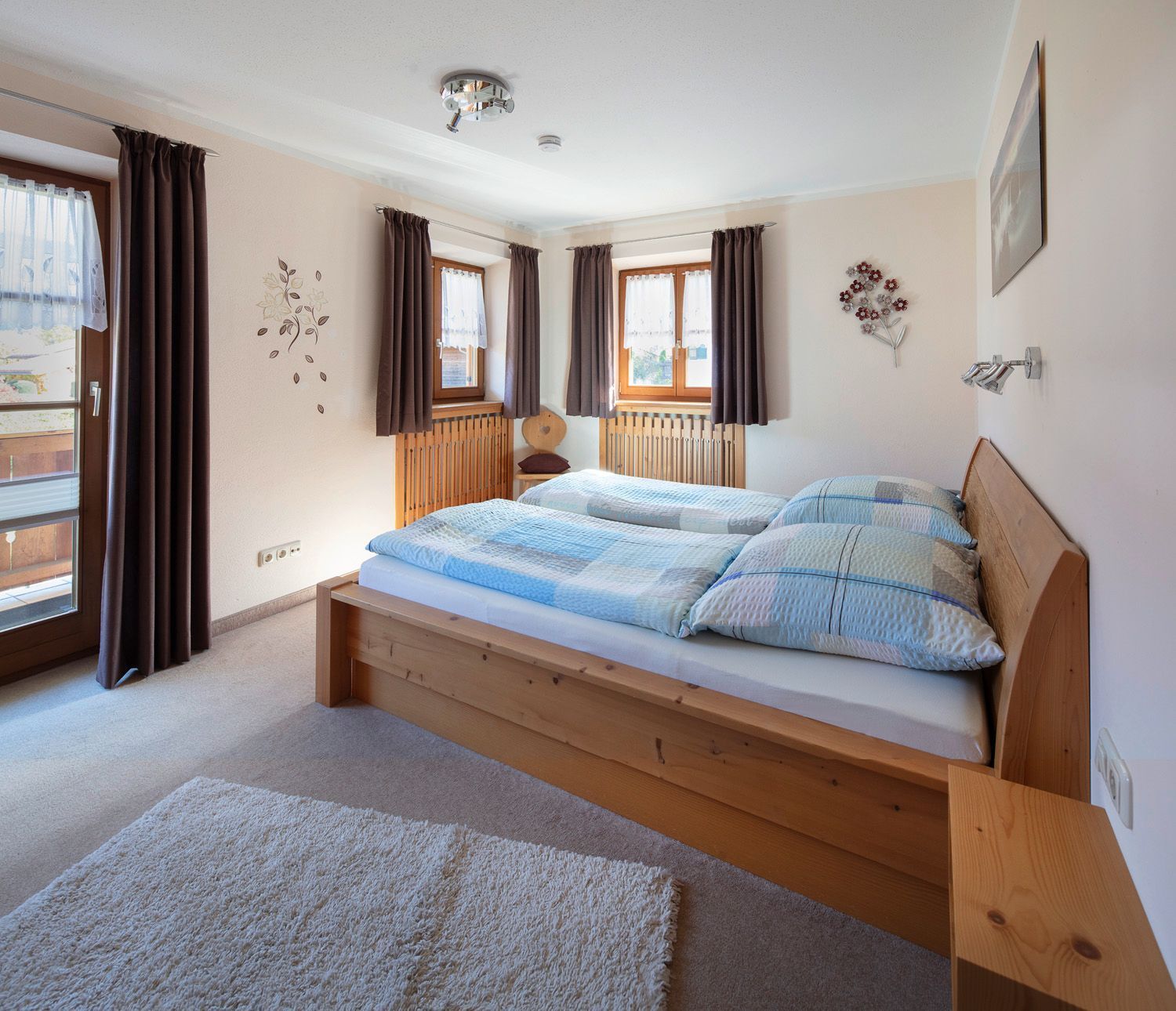 Ein Schlafzimmer mit einem hölzernen Bett, zwei Fenstern mit Vorhängen, einem Heizkörper und einer Wandleuchte.