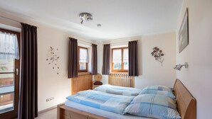 A bedroom with a wooden bed, two windows with curtains, a radiator, and a wall-mounted light fixture. - Apartment 3 - Apartments Lohbinder (Fam. Holzer) (Rottach-Egern)