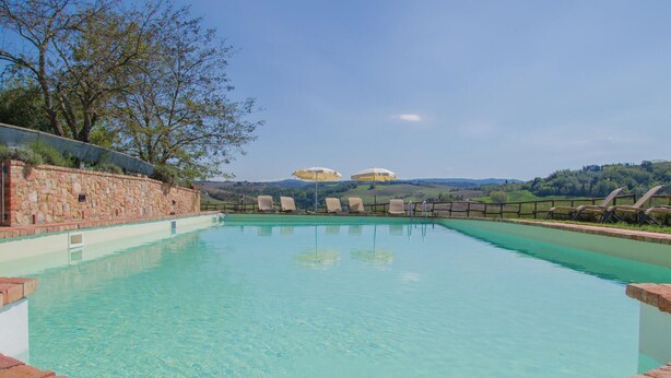 Una piscina amb aigua cristal·lina, envoltada per una paret de pedra i una tanca de fusta. Hi ha gandules i ombrelles al costat de la piscina.