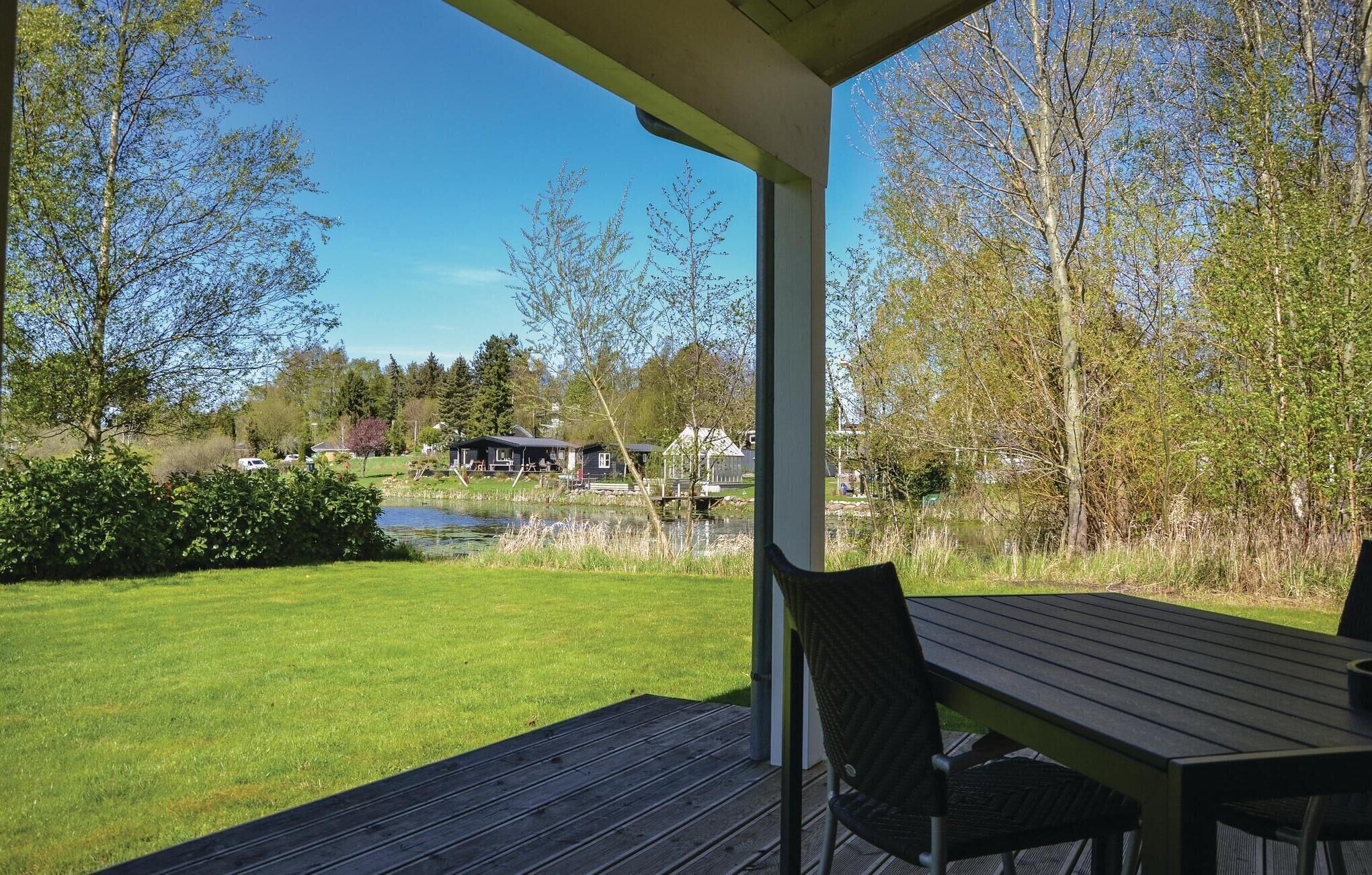 Ein überdachter Außenbereich mit Tisch und Stühlen, mit Blick auf einen Teich und eine Rasenfläche mit Bäumen und Häusern im Hintergrund.
