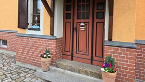 A building with a red door, a window with brown shutters, and two flower pots on the steps. - Single Room - Pension Max (Senftenberg)