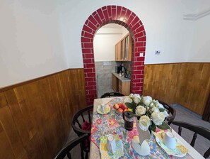 Un comedor con una mesa lista para dos personas, con mantel estampado con flores, cafetera y un florero con flores blancas.