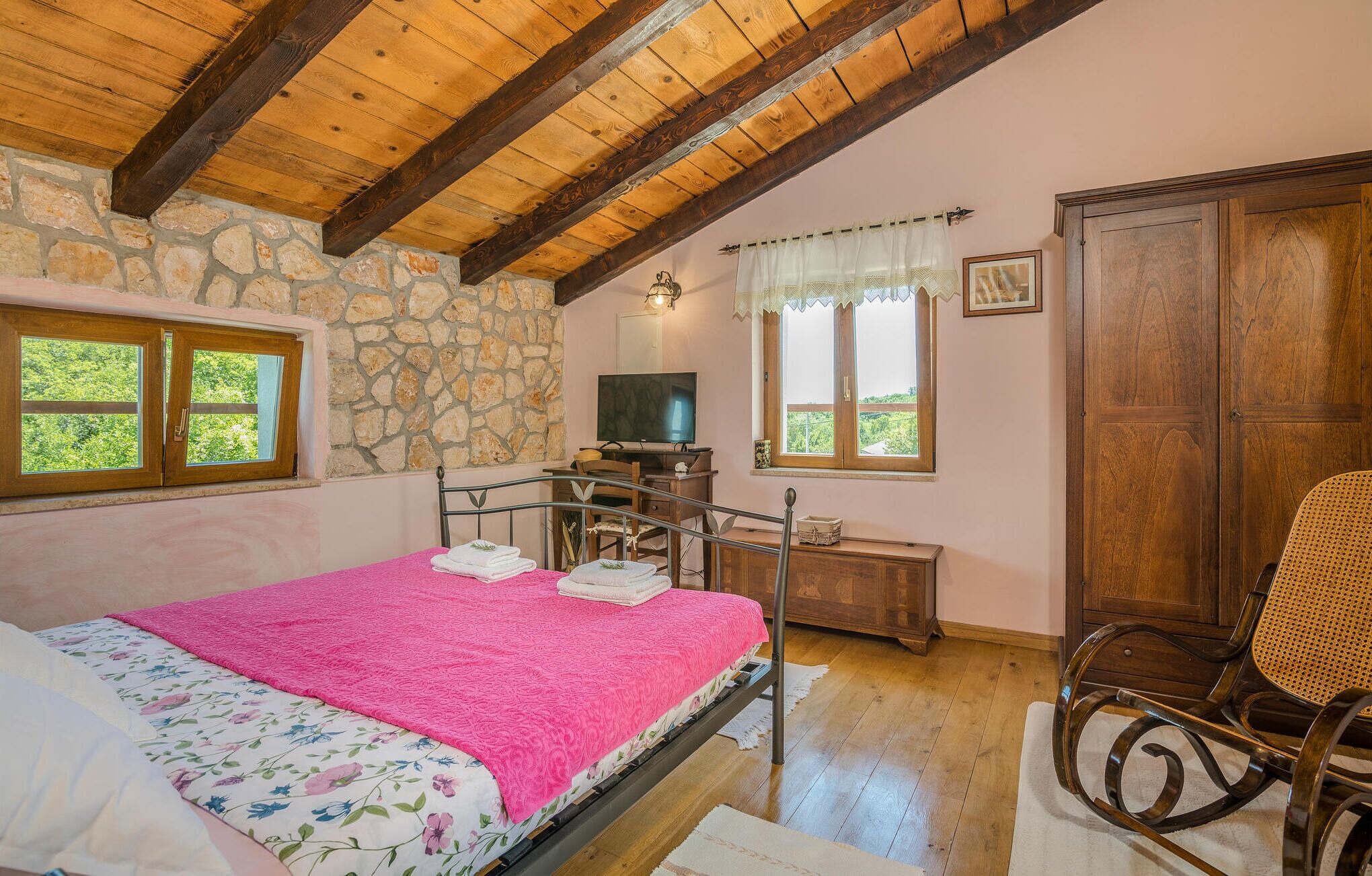 Ein Schlafzimmer mit Steinwand, Holzbalkendecke, einem Bett mit rosa Decke, einem Fernseher, einem Holzschrank und einem Fenster mit Blick.