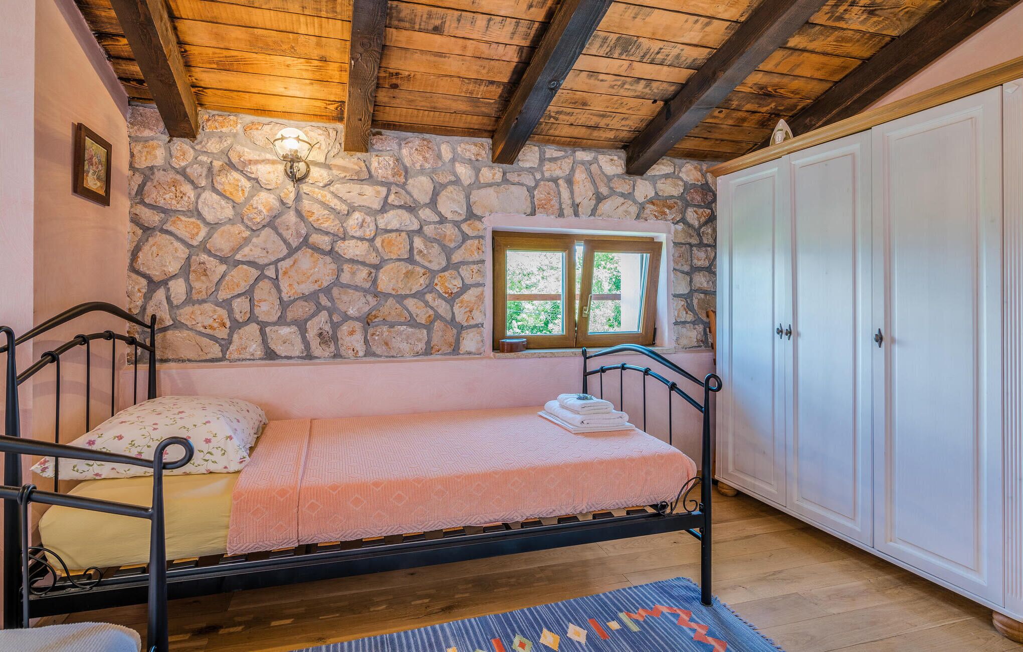 Un dormitorio con pared de piedra, una cama con manta rosa, una ventana y un armario.