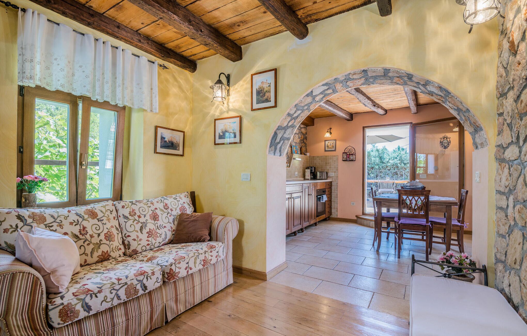 A cozy living room with a floral sofa, a dining area with a table and chairs, and a kitchen with stone archway.