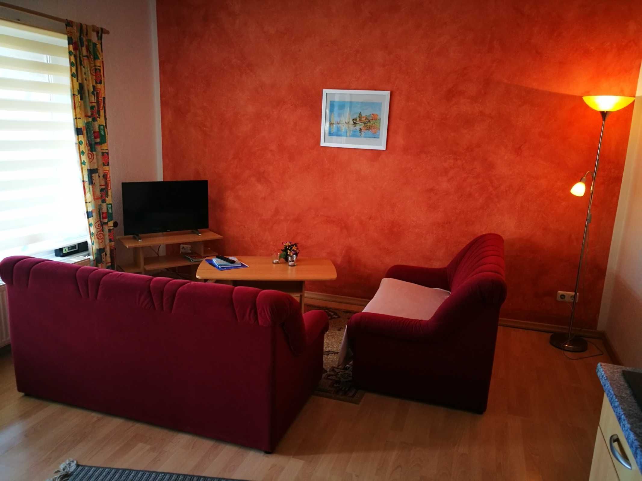Un salon avec un canapé rouge, une petite table sur laquelle se trouve une télécommande, un téléviseur et une lampe.