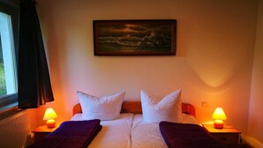 Un lit double avec du linge de lit blanc, deux oreillers blancs et un plaid rouge foncé sur chaque côté. Il y a deux tables de chevet avec des lampes, et un tableau représentant un paysage marin est accroché au-dessus du lit.