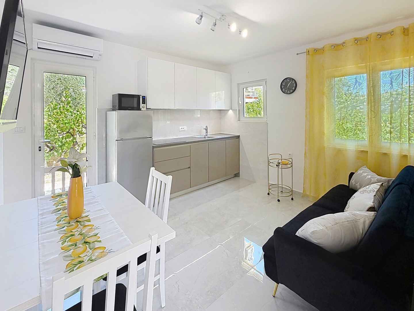 A compact kitchen with white cabinets, a microwave, and a sink. There's a dining table with chairs and a black sofa with cushions.