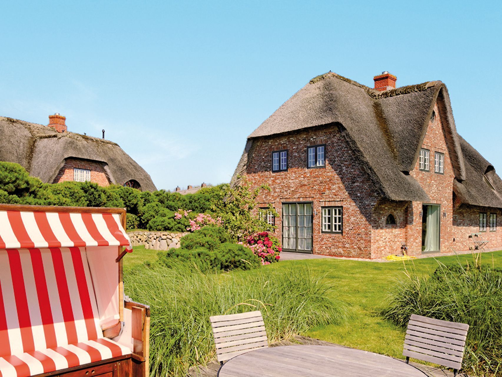 A traditional thatched-roof house with a brick lower section, surrounded by lush greenery and a wooden deck with a striped chair.