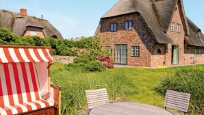 A traditional thatched-roof house with a brick lower section, surrounded by lush greenery and a wooden deck with a striped chair. - House to the lighthouse by A&M - House to the lighthouse (Kampen (Sylt))