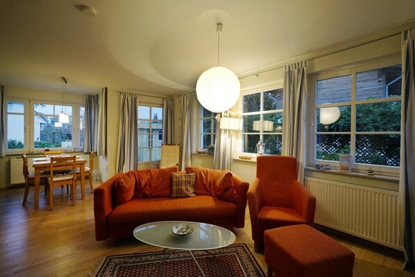 A living room with a sofa, armchair, dining table, and chairs, large windows with curtains, and a hanging light fixture.