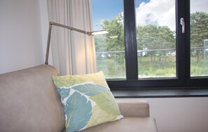 Ein Zimmer mit einem beigefarbenen Sofa, einem gemusterten Kissen, einer Stehlampe und einem Fenster mit Blick auf Bäume und einen Zaun.