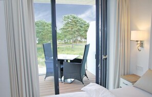 A hotel room with a bed, a small bedside table, a lamp, a balcony with a table and chairs, and a view of trees and grass.