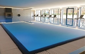 An indoor swimming pool with clear glass walls, a tiled floor, and a few lounge chairs along the side.