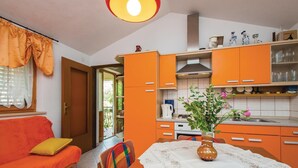 Une cuisine de taille réduite avec des meubles de couleur orange, une table à manger recouverte d’une nappe blanche et un canapé agrémenté d’un coussin jaune.
