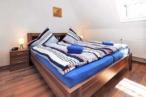 A neatly made bed with a striped blanket and pillows, a bedside table with a lamp, and a window with blinds. - Holiday home beach chair (Esens)