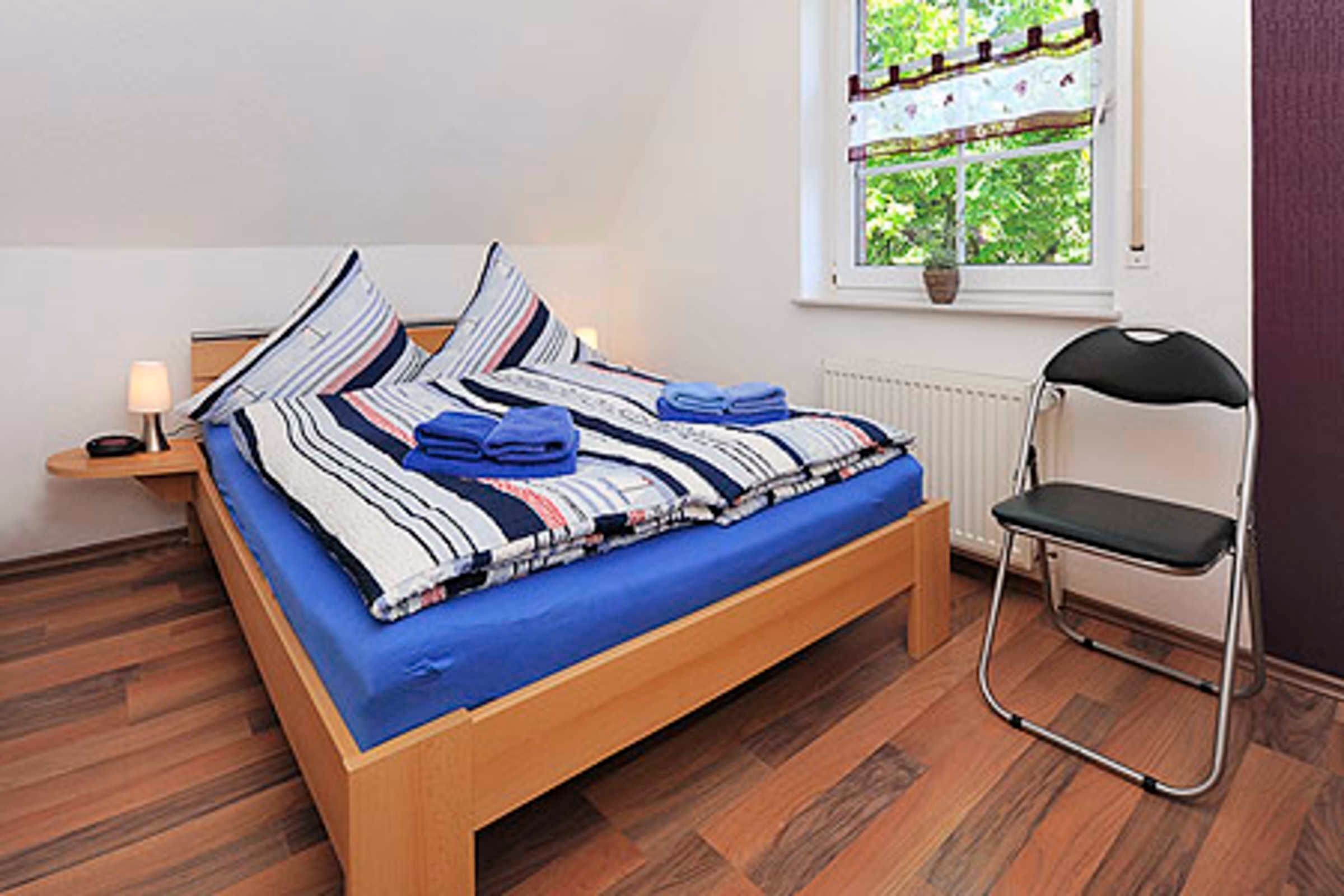 Una cama con colchón azul, ropa de cama a rayas y toallas dobladas. Hay una silla junto a la cama y una ventana con vistas a la vegetación.