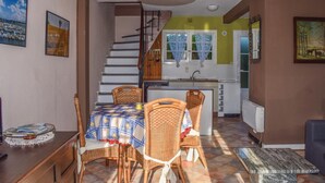 Un coin repas avec des chaises en osier, une table recouverte d’une nappe à carreaux et un escalier menant à la cuisine.