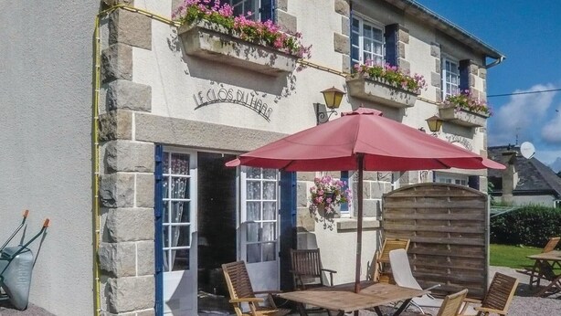 Un bâtiment en pierre avec un parasol rouge, des tables à l’extérieur et des jardinières fleuries.