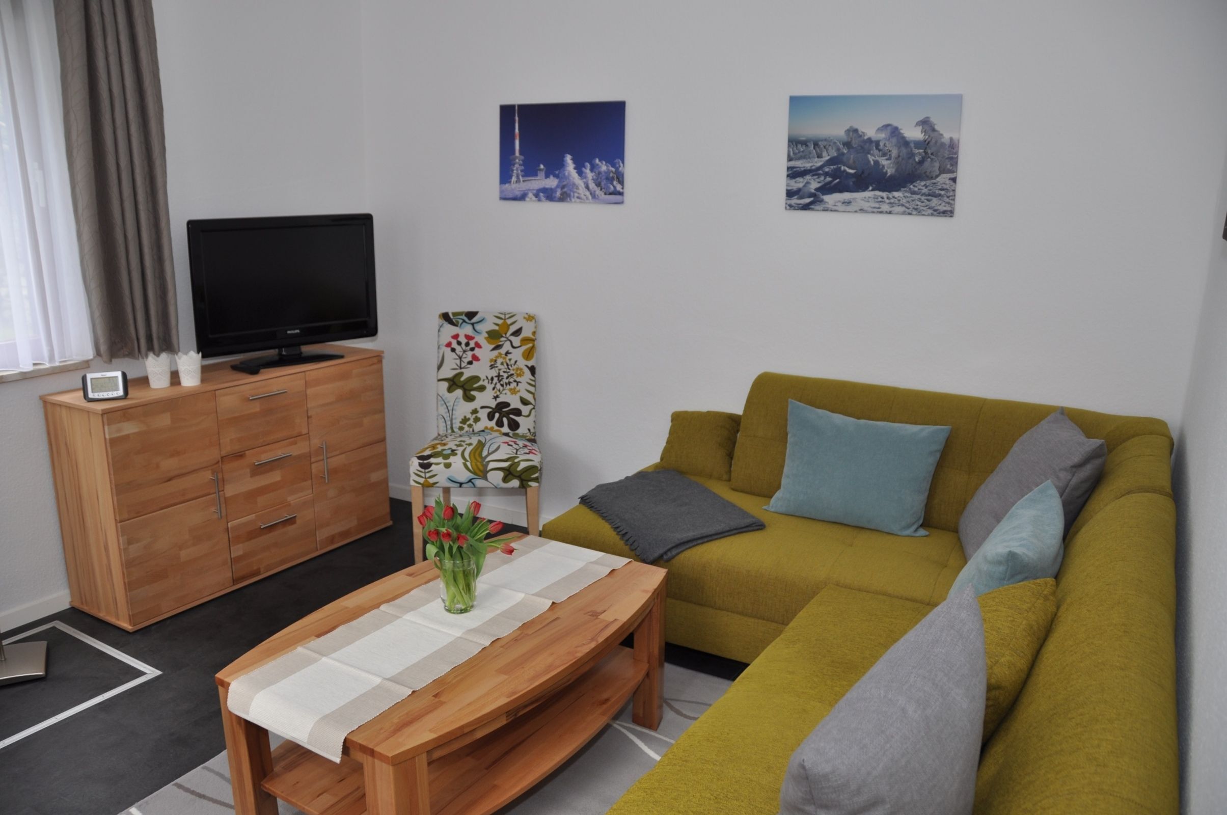 Uma sala de estar com um sofá, uma mesa de centro de madeira, uma televisão em um armário e duas imagens penduradas na parede.