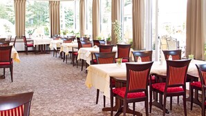 A dining area with multiple tables and chairs, large windows with curtains, and a carpeted floor.