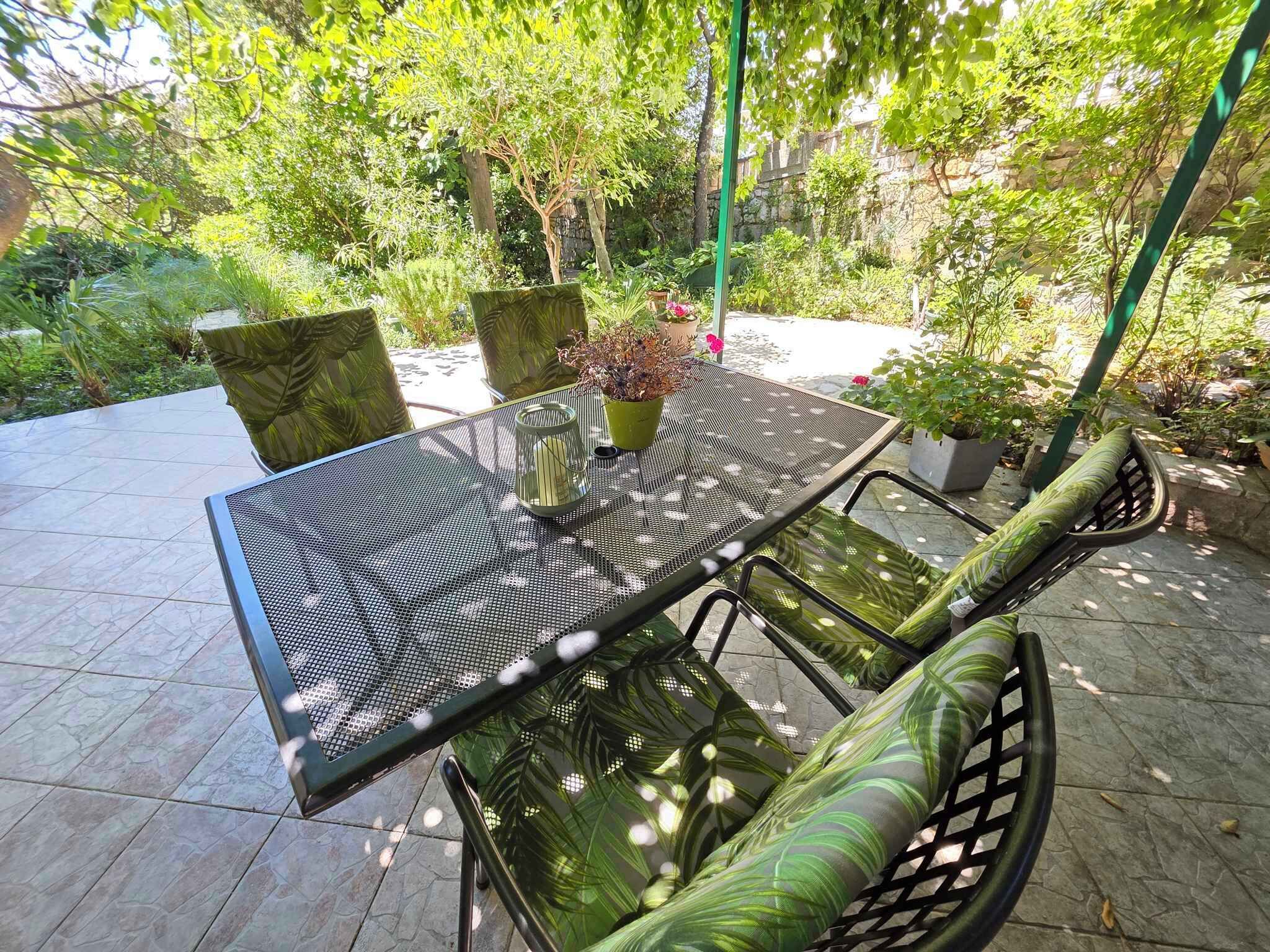 Eine Terrasse im Freien mit einem Metalltisch und -stühlen, umgeben von Grünpflanzen und Topfpflanzen.