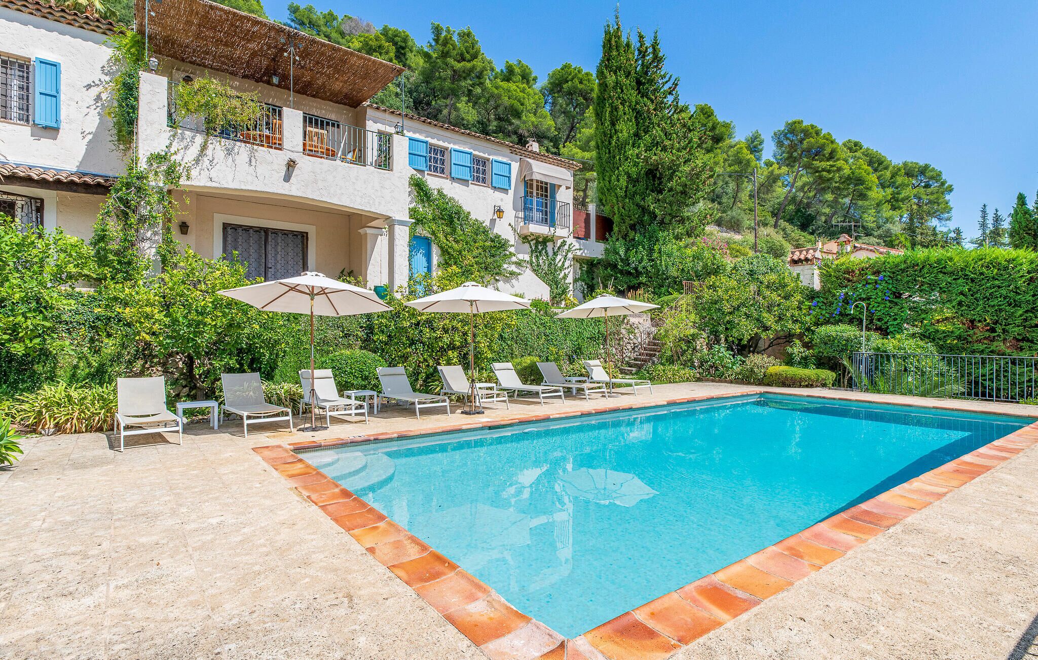 A swimming pool with lounge chairs and umbrellas, surrounded by lush greenery and a multi-story building with balconies.