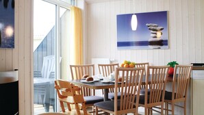 Un coin repas avec des chaises en bois et une table dressée pour le petit-déjeuner. On y voit un tableau représentant une bougie sur une table, ainsi qu’une fenêtre avec des rideaux.